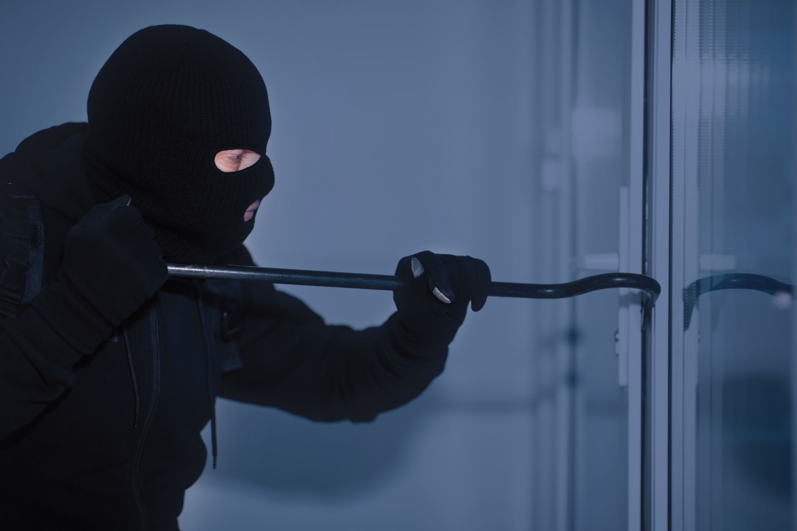 Robber in black balaclava cracking door with crowbar